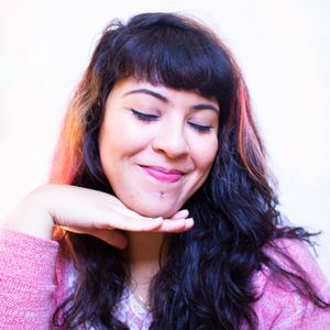 Selfie de Camila Marciano. Mulher negra de pele clara com cabelos longos da cor preta e mechas pintadas em tons de laranja e rosa, fazendo pose com a mão embaixo do queixo.